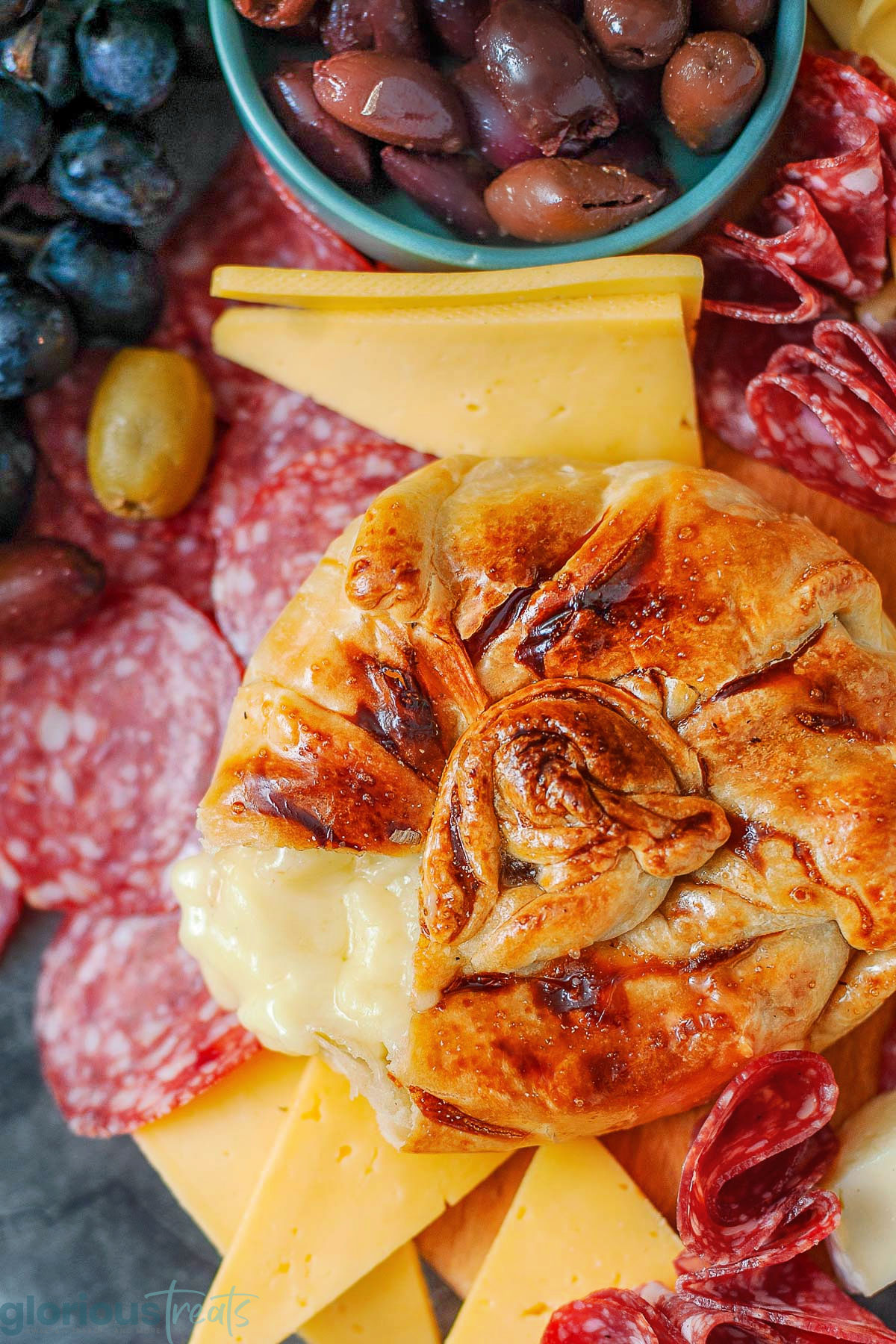 Honey baked brie wrapped in puff pastry. Salami, cheese, olives and grapes are on the platter with the brie.