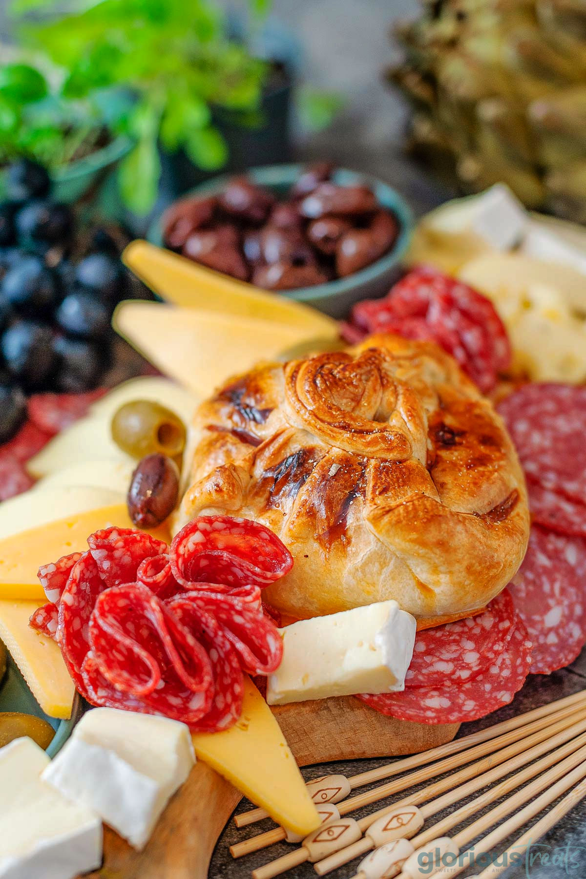 Honey baked brie wrapped in puff pastry. Salami, cheese, olives and grapes are on the platter with the brie.