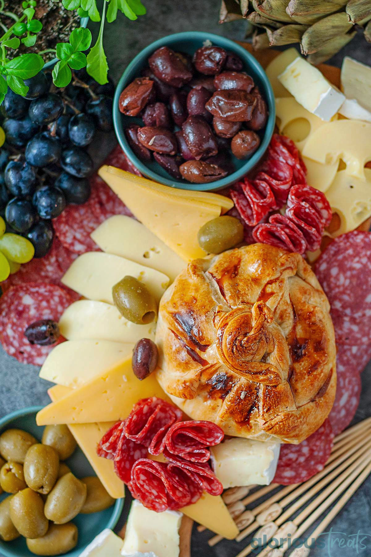 Honey baked brie wrapped in puff pastry. Salami, cheese, olives and grapes are on the platter with the brie.