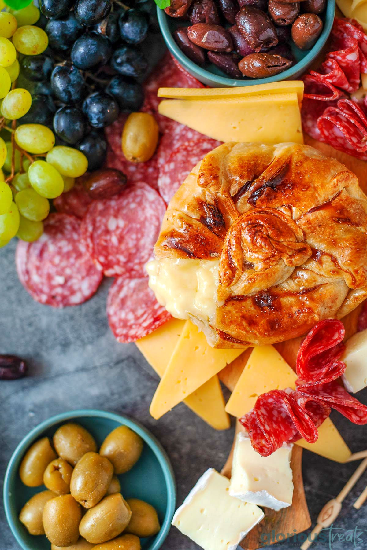 Honey baked brie wrapped in puff pastry with a large scoop taken out of it. Salami, cheese, olives and grapes are on the platter with the brie.