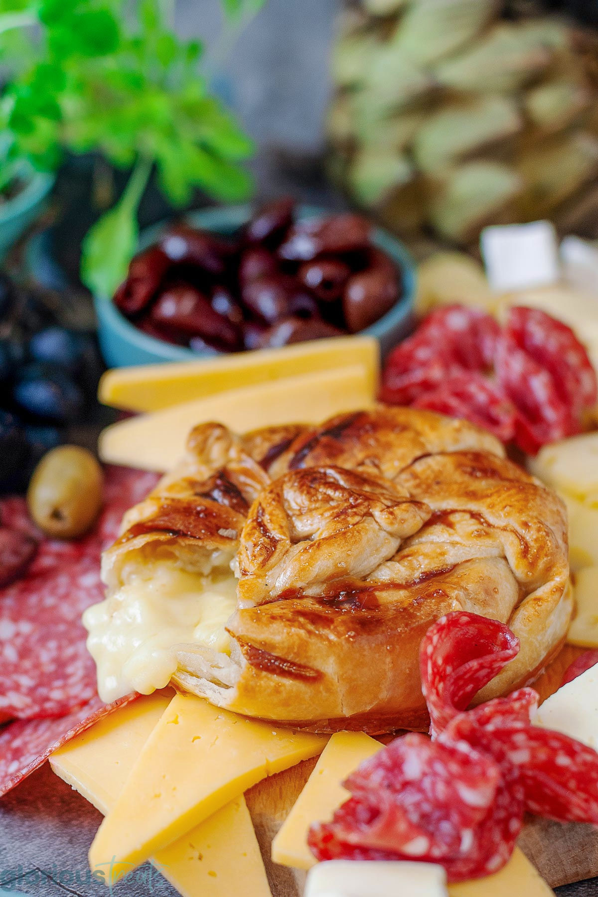 Honey baked brie wrapped in puff pastry with a large scoop taken out of it. Salami, cheese, olives and grapes are on the platter with the brie.