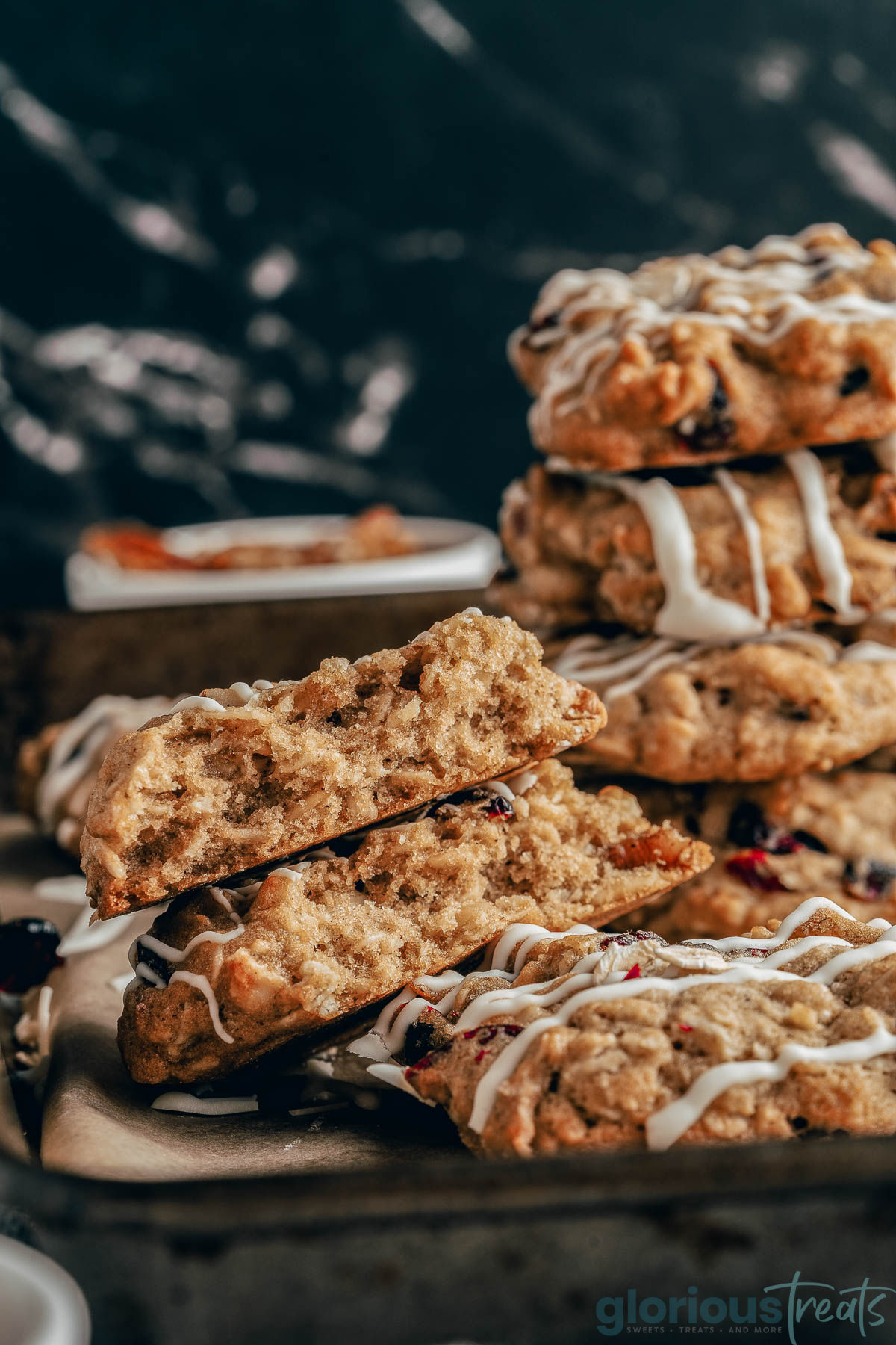 A close up view of cranberry oatmeal cookies on a baking sheet. One cookie is broken in half and several more are stacked in the background.