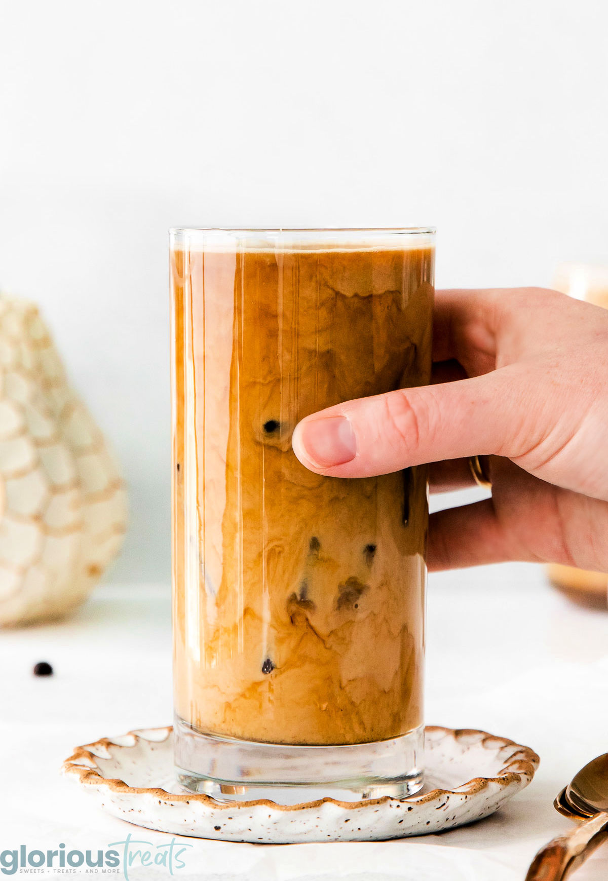 A side view of a hand holding a glass of pumpkin cream cold brew. A decorative plate is underneath the glass.