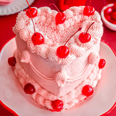 Heart shaped cherry cake recipe decorated with cherry frosting and maraschino cherries on cake stand.