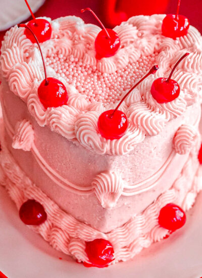 Heart shaped cherry cake recipe decorated with cherry frosting and maraschino cherries on cake stand.