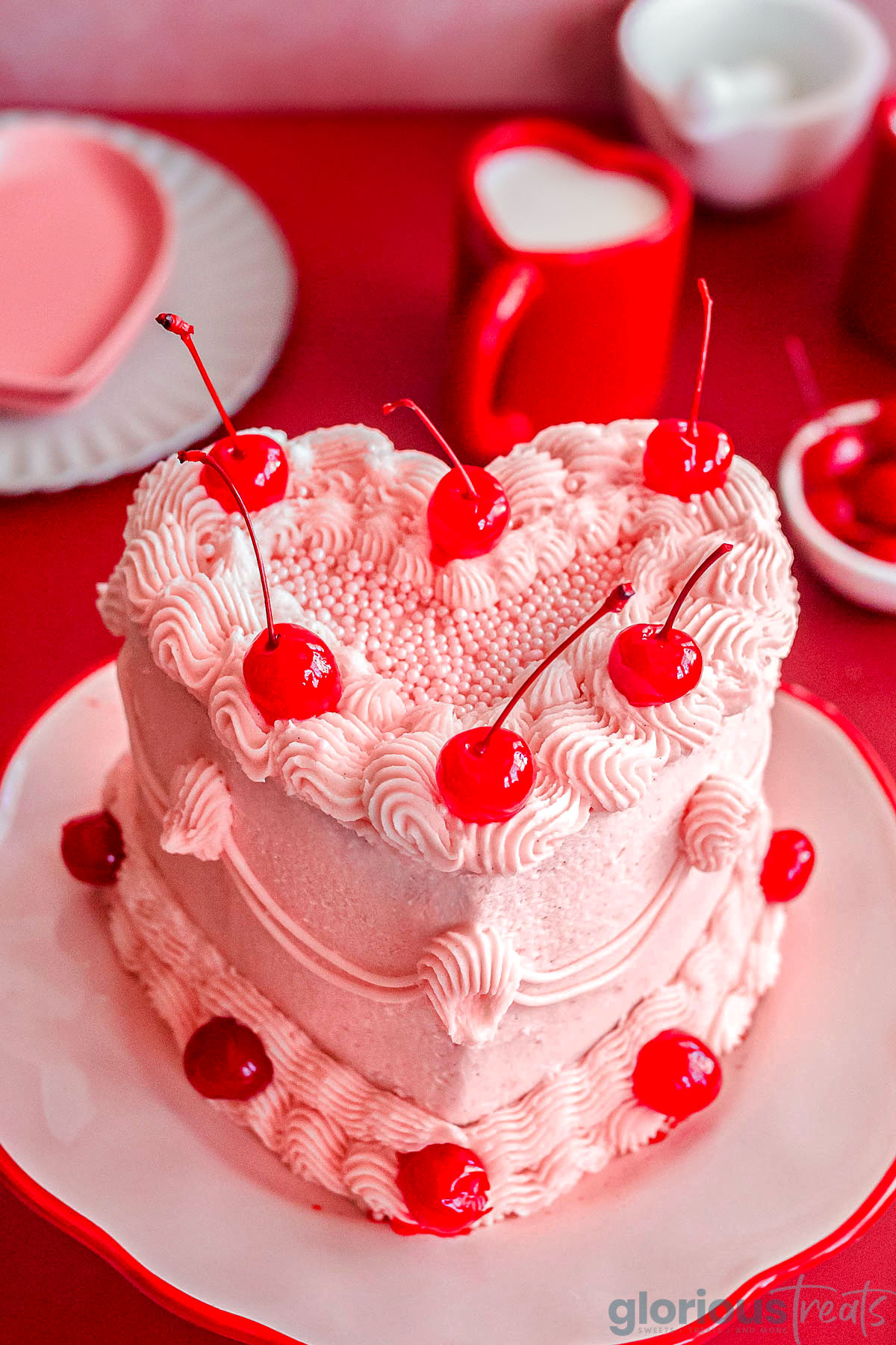 Heart shaped cherry cake recipe decorated with cherry frosting and maraschino cherries on cake stand.