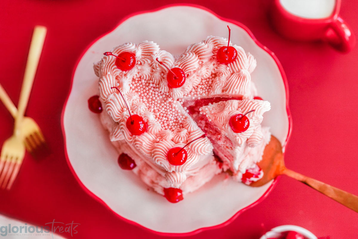 Top down look at cherry chip cake decorated with cherries. Slice of cake is being cut from the rest of the cake and removed with a server.