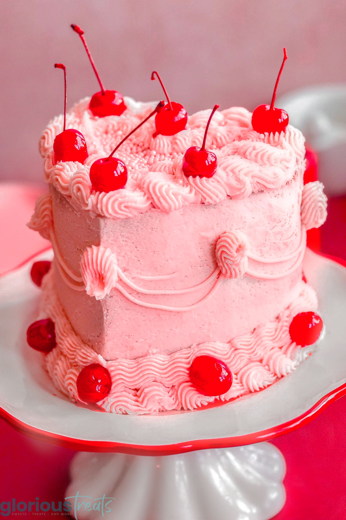 Heart shaped cherry cake recipe decorated with cherry frosting and maraschino cherries on cake stand.