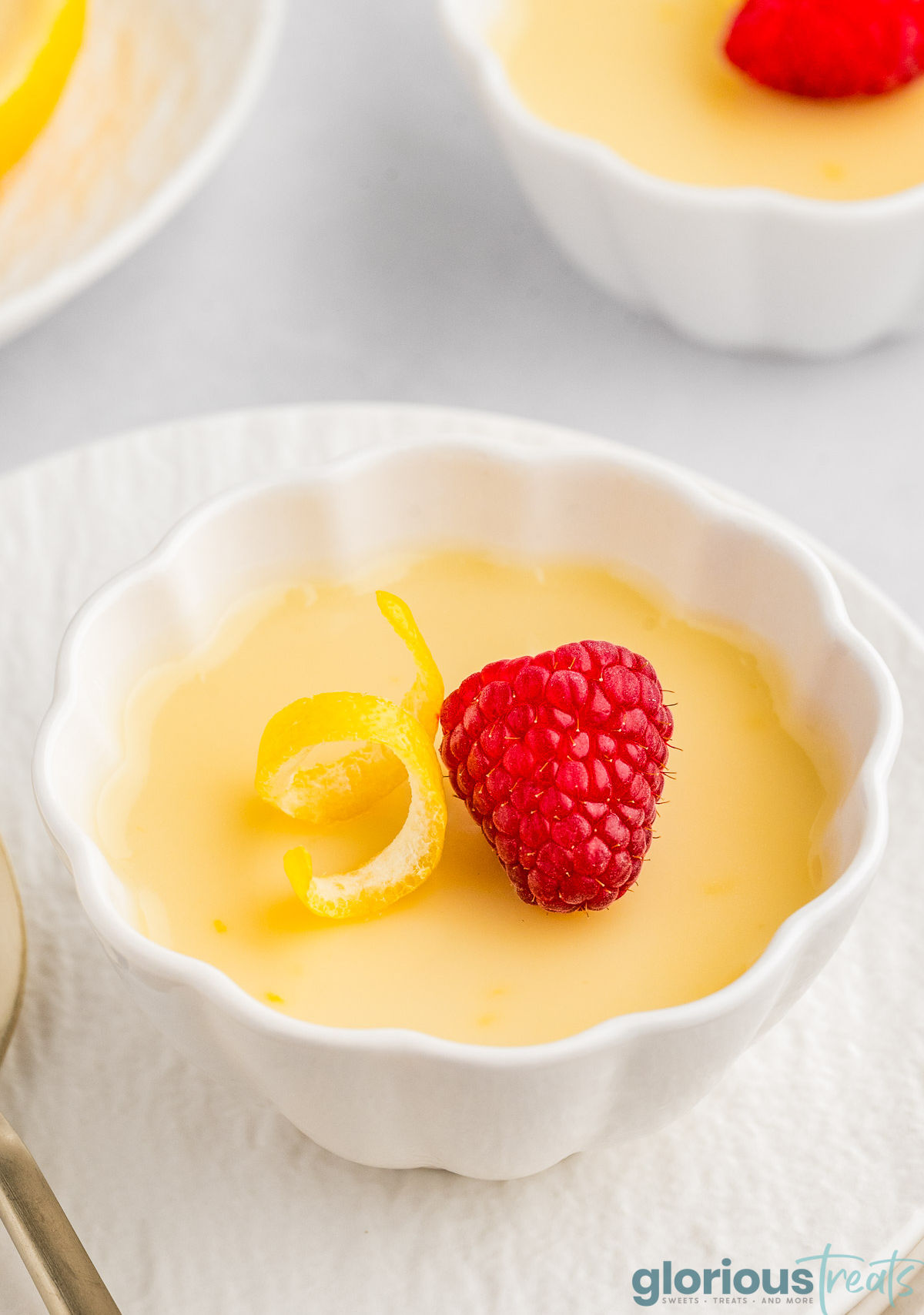 A close up view of lemon posset in a white decorative bowl on a plate. More posset is in the background.